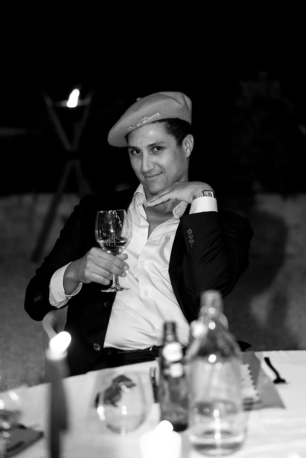 A man in formal attire holding a glass of wine at an outdoor dinner party at night, wearing a beret tilted to one side.