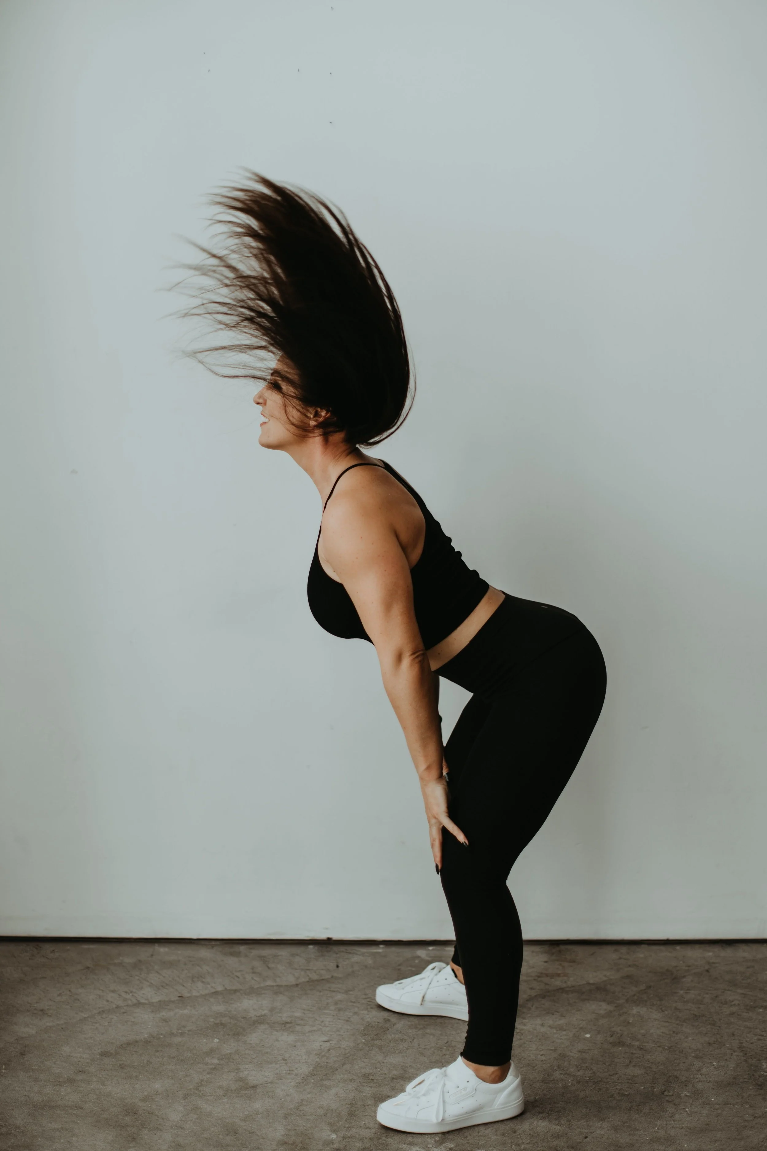 Woman with long hair and light skin in black athletic clothes, leaning forward with hair flying, on a concrete floor against a plain light background.