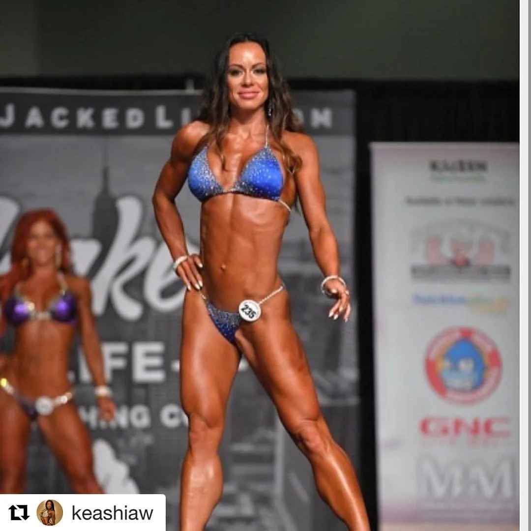 A female bodybuilder on stage during a competition, wearing a sparkly blue bikini, posing with one hand on her hip, with a background banner and another competitor behind her.