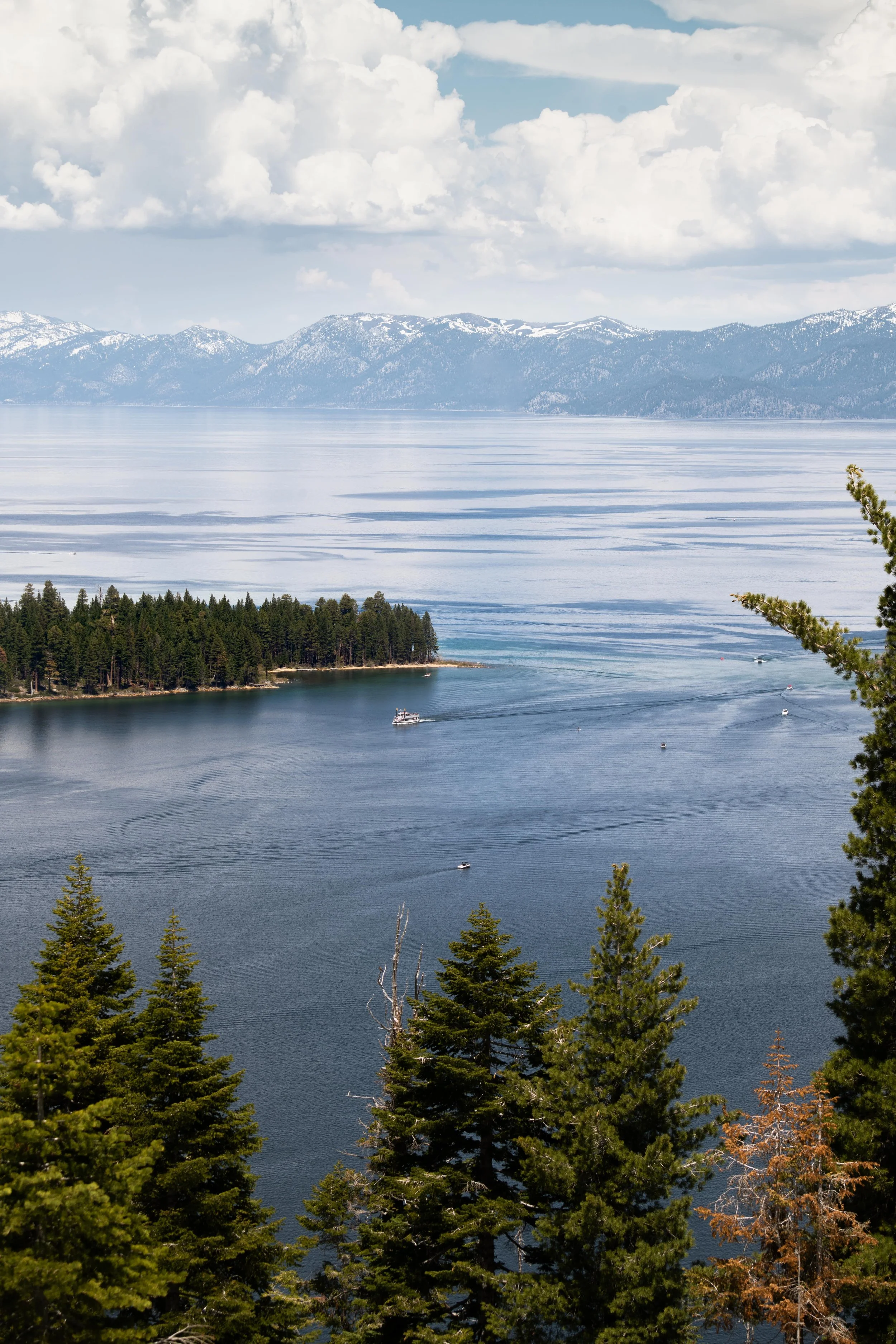 A scenic view of a Lake Tahoe from Emerald Bay with surrounded mountains, a forested shoreline, boats on the water, and partly cloudy sky.