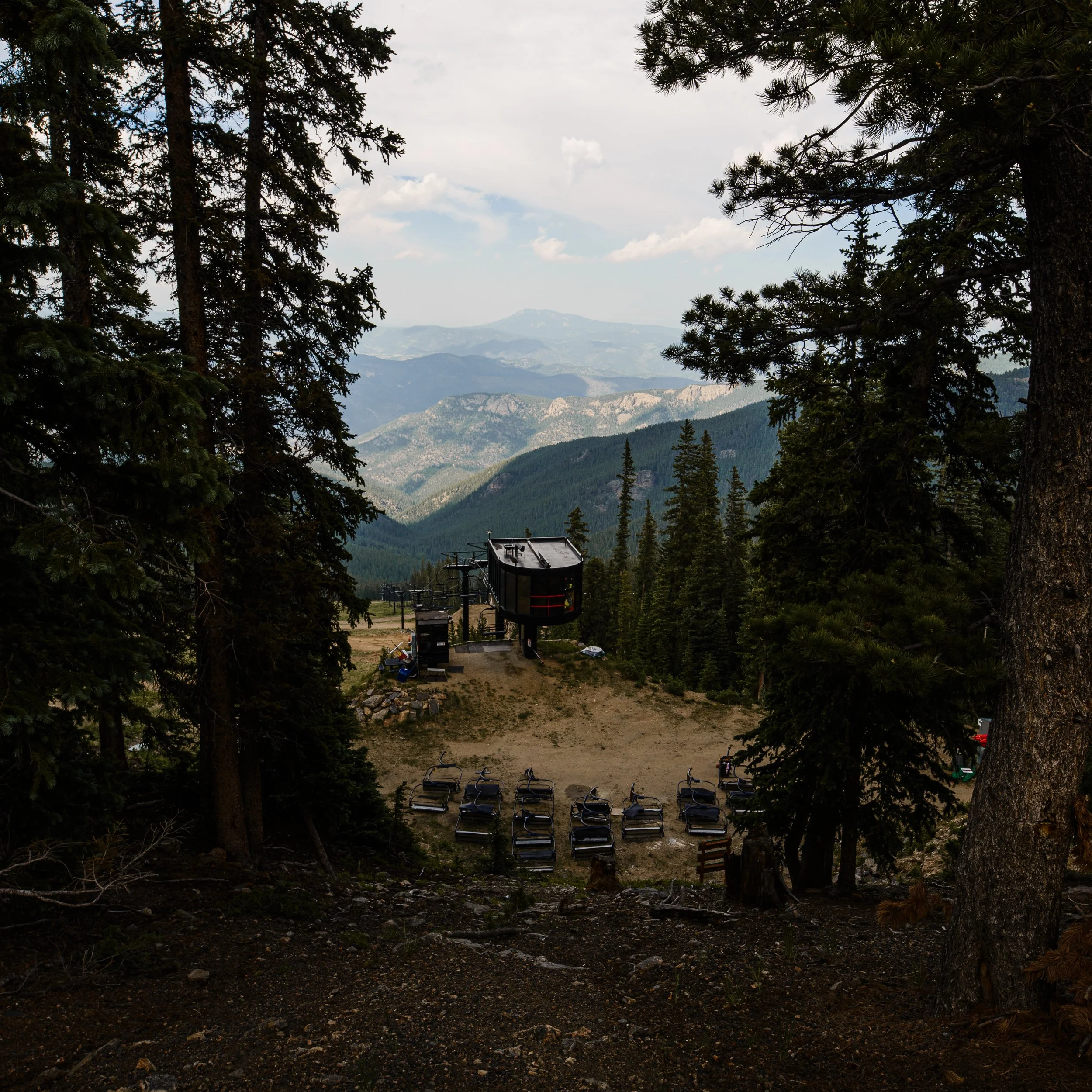 Mountain landscape with dense pine forest, ski lift equipment, and empty ski chairs in the foreground. Distant mountain ranges under partly cloudy sky.
