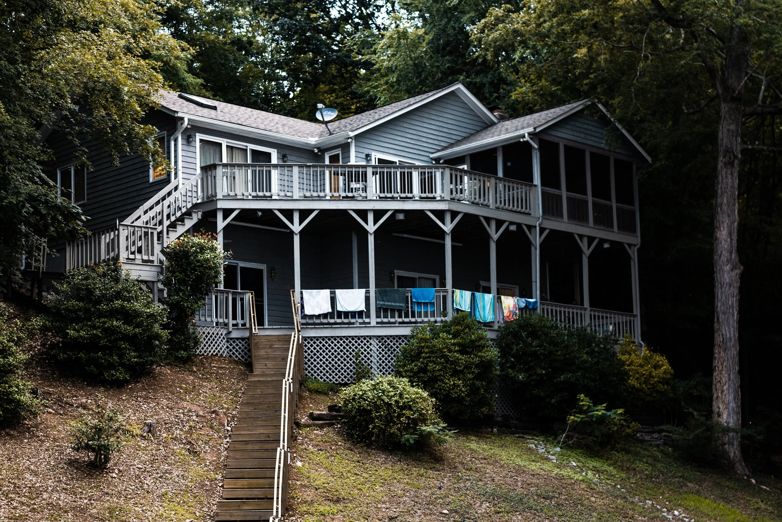 A two-story lakehouse with surrounding trees, featuring a large wooden deck with laundry hanging to dry, and stairs leading up from a sloped yard.