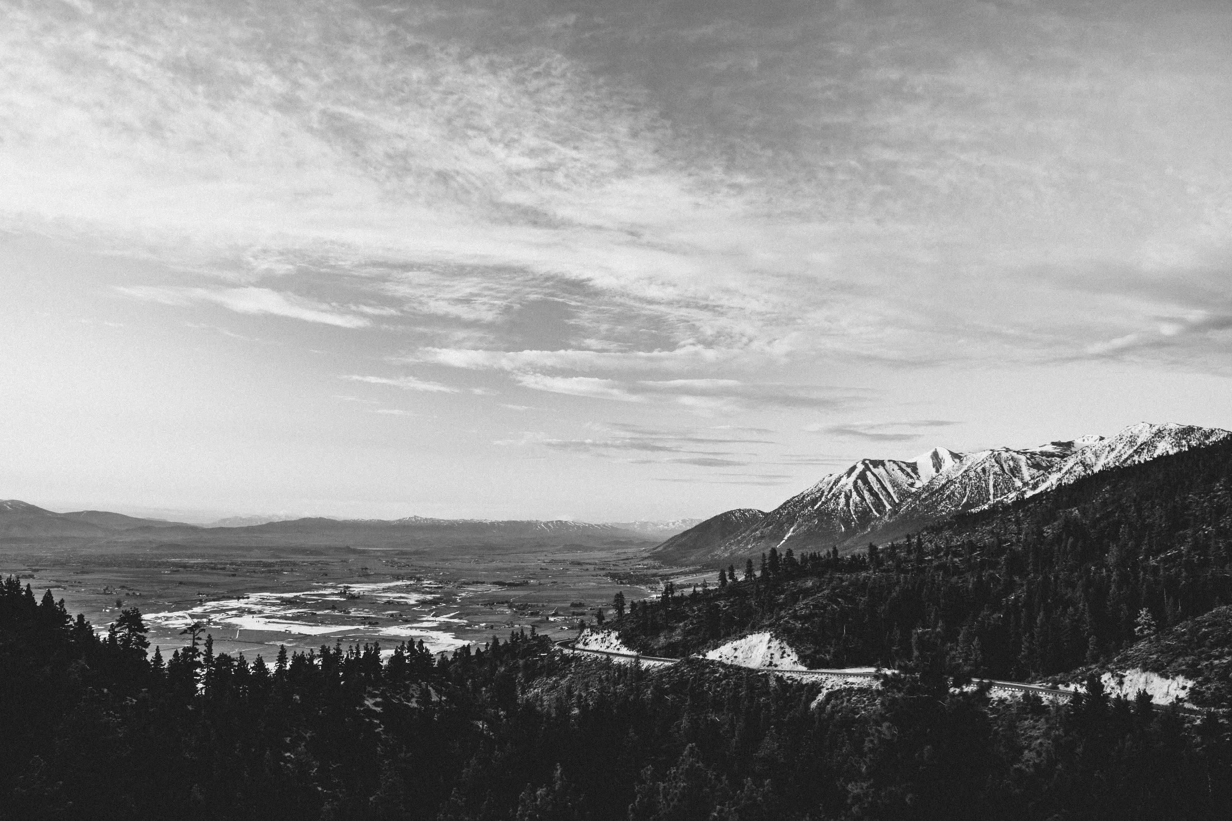 Black and white landscape of mountains with snow, forests, and a valley with fields and roads under a cloudy sky.