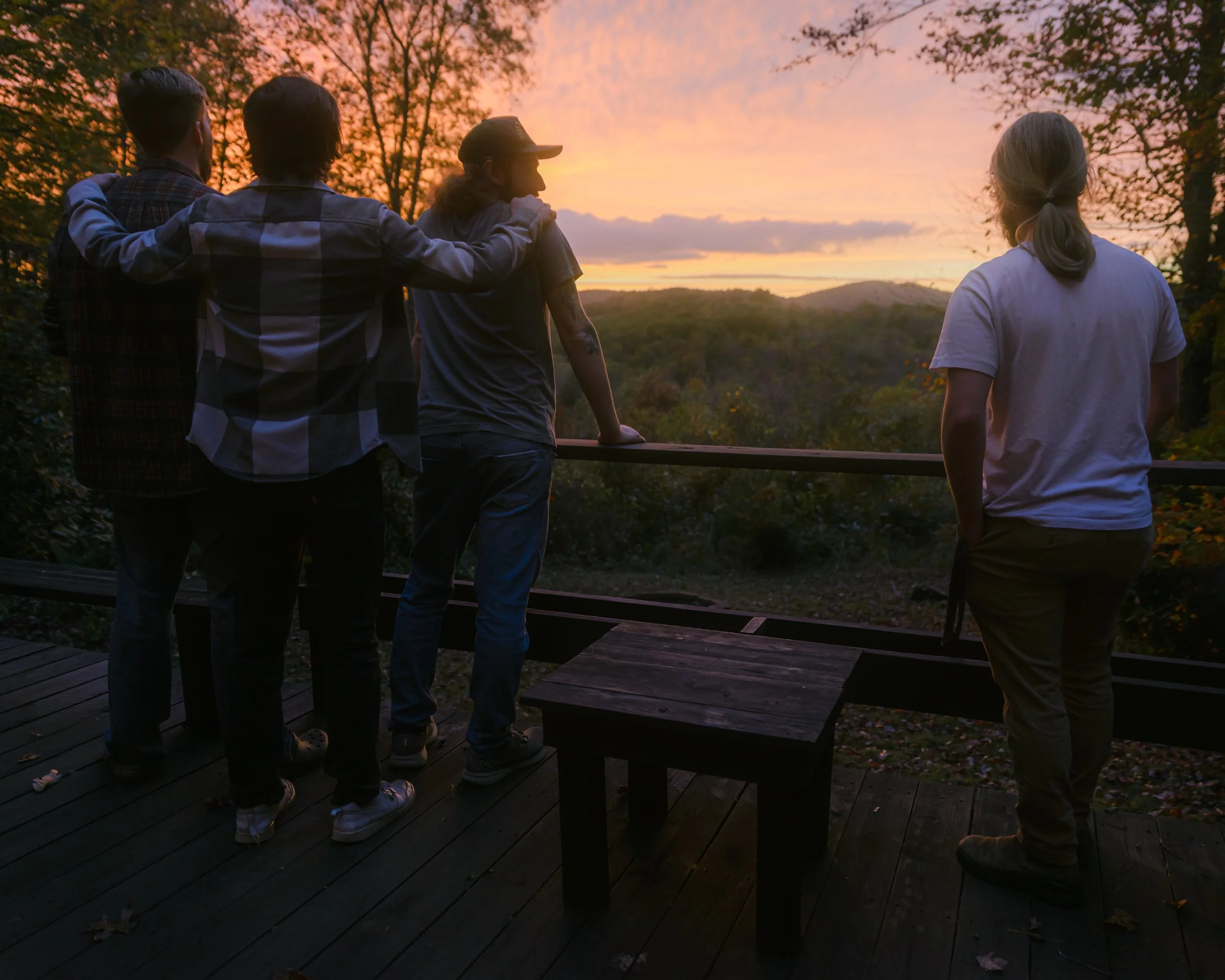Four people standing on a wooden deck, overlooking a sunset landscape with trees and hills, some with arms around each other.