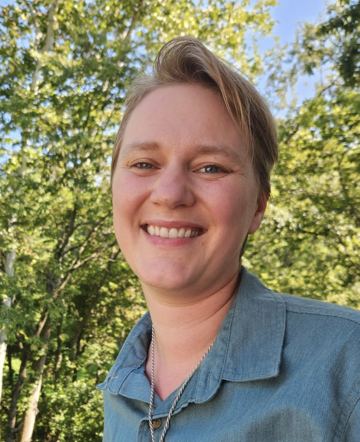 A smiling person with short blond hair, wearing a denim shirt and a necklace, standing outdoors with a background of green trees and a blue sky.