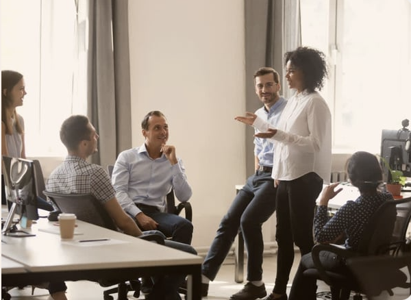 A diverse group of six people in an office, engaged in a discussion. One woman is standing and speaking, while four others are seated around a table, listening and smiling. The office has large windows with curtains and a computer on the desk.