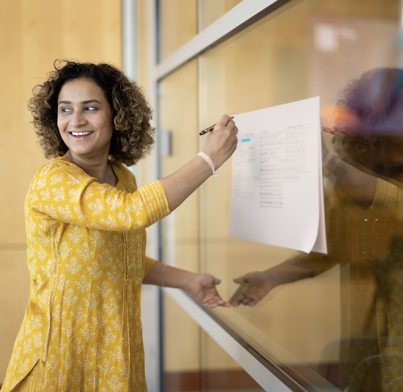 A woman with curly hair wearing a yellow dress with white patterns, writing on a paper attached to a glass board in a classroom or office setting.