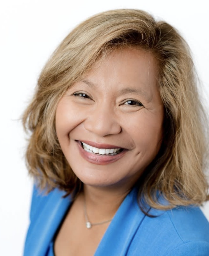 A smiling woman with shoulder-length blonde hair, wearing a blue blazer and a necklace, against a white background.