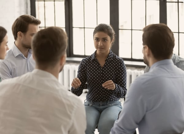 A young woman moderates a group discussion in an office setting with large windows behind her.
