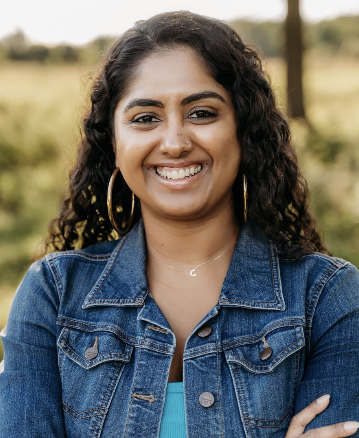 Smiling woman with dark curly hair, wearing large hoop earrings, a denim jacket, and a turquoise top, outdoors in a natural setting.