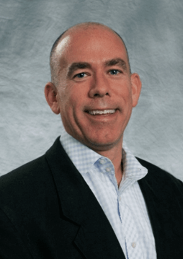 Headshot of a smiling man with a bald head, wearing a dark blazer and a light-colored dress shirt against a gray background.
