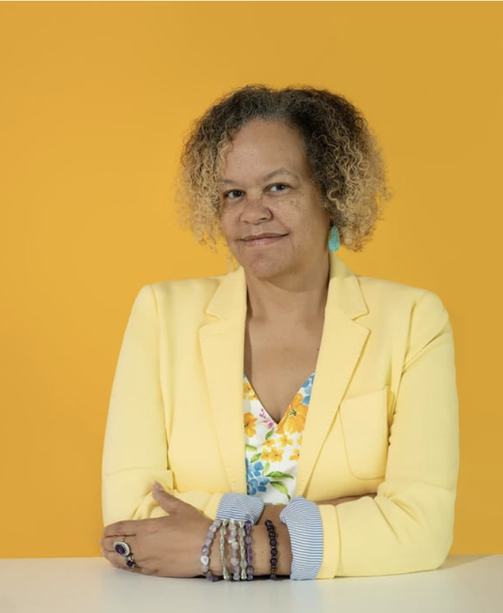 A woman with curly blonde hair wearing a yellow blazer and floral top, sitting at a white table with her arms crossed, against a yellow background.