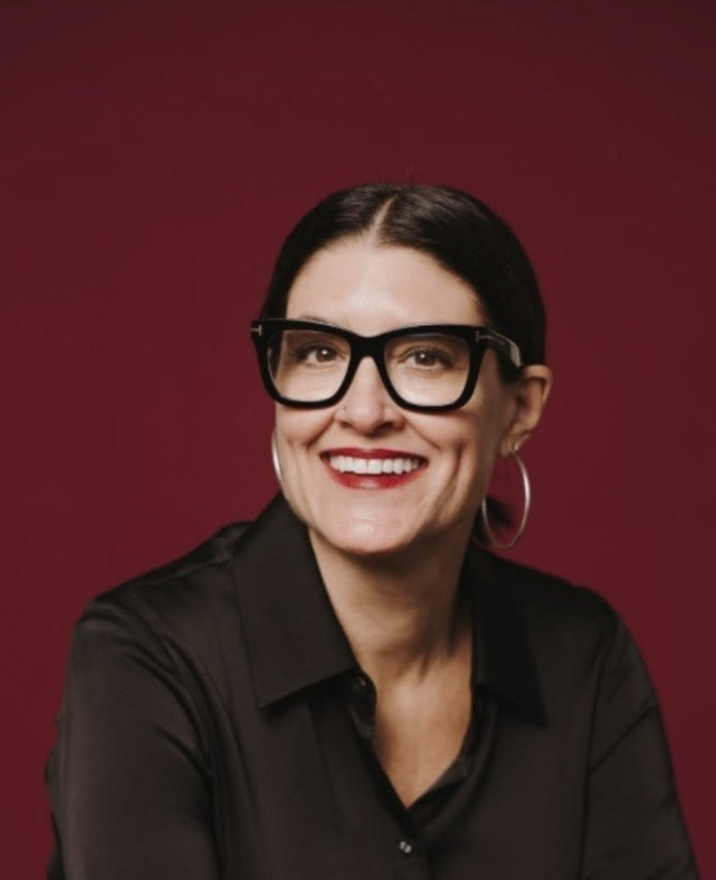 A woman with dark hair, wearing large black glasses, earrings, a black shirt, and red lipstick, smiling against a maroon background.