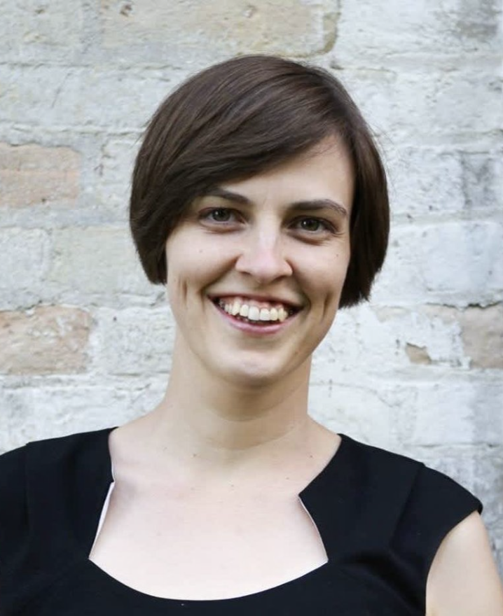 A woman with short brown hair, smiling, wearing a black top, standing in front of a light gray brick wall.