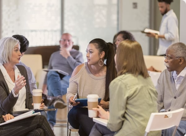 A group of diverse people sitting in a circle during a discussion in a room with large windows, with a man standing taking notes.