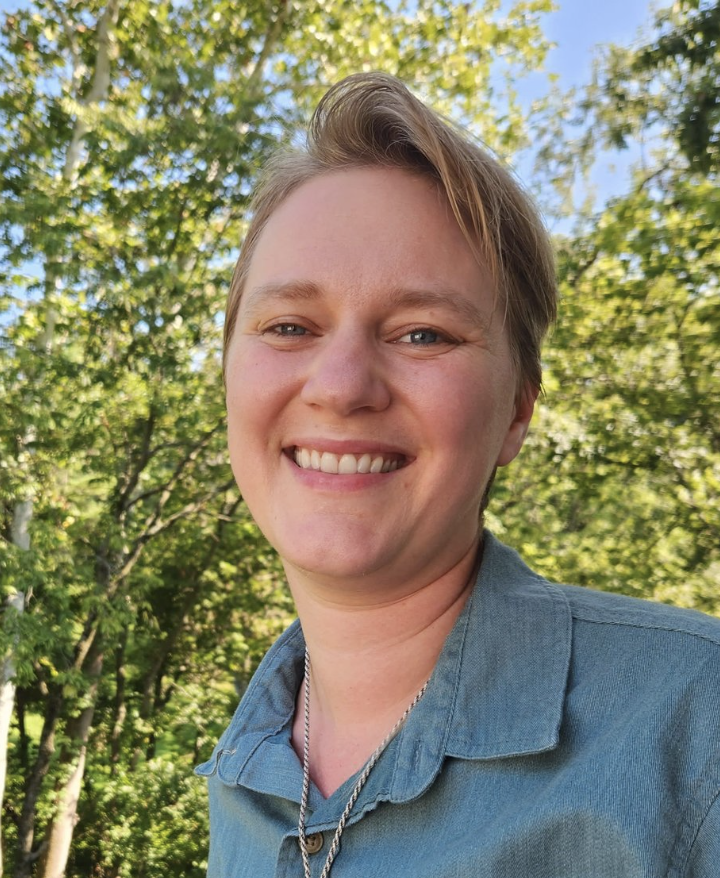 A person smiling outdoors with trees and blue sky in the background.