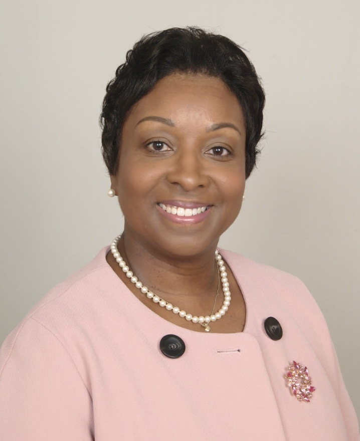 A smiling woman with short dark hair wearing a pink blazer, pearl necklace, pearl earrings, and a decorative brooch, standing against a plain background.