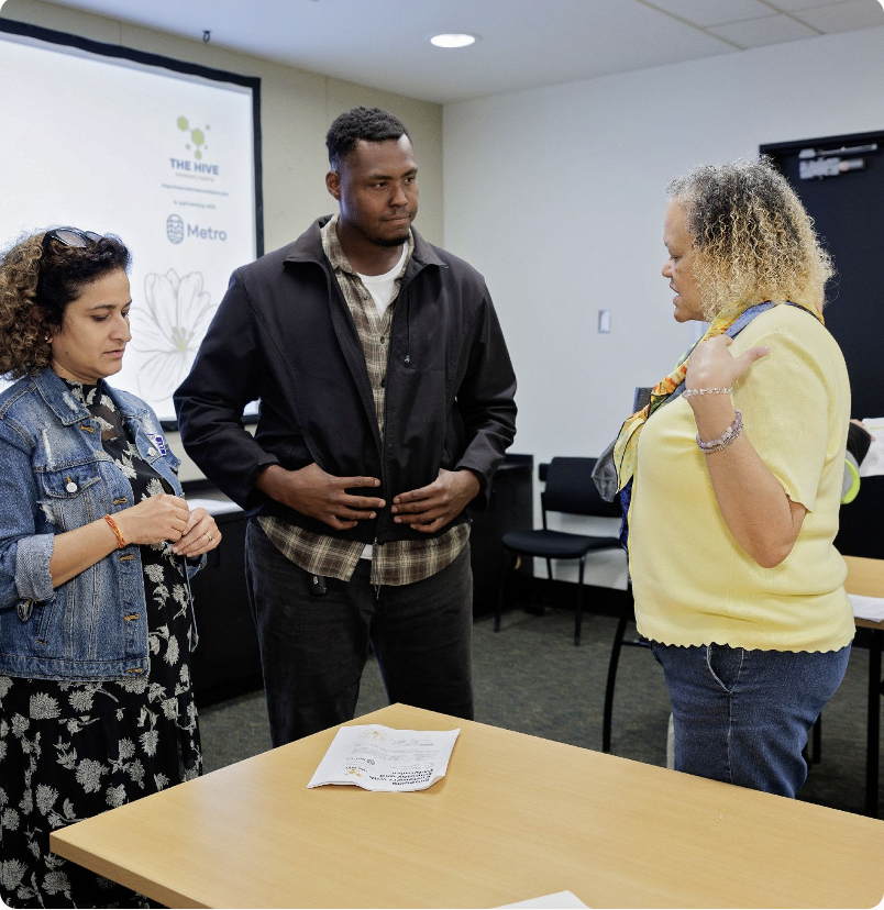 Three people standing and talking in a conference room, with a table and chairs around. A woman in a yellow shirt appears to be explaining or talking, while two others listen. There is a screen in the background with a logo and some text.
