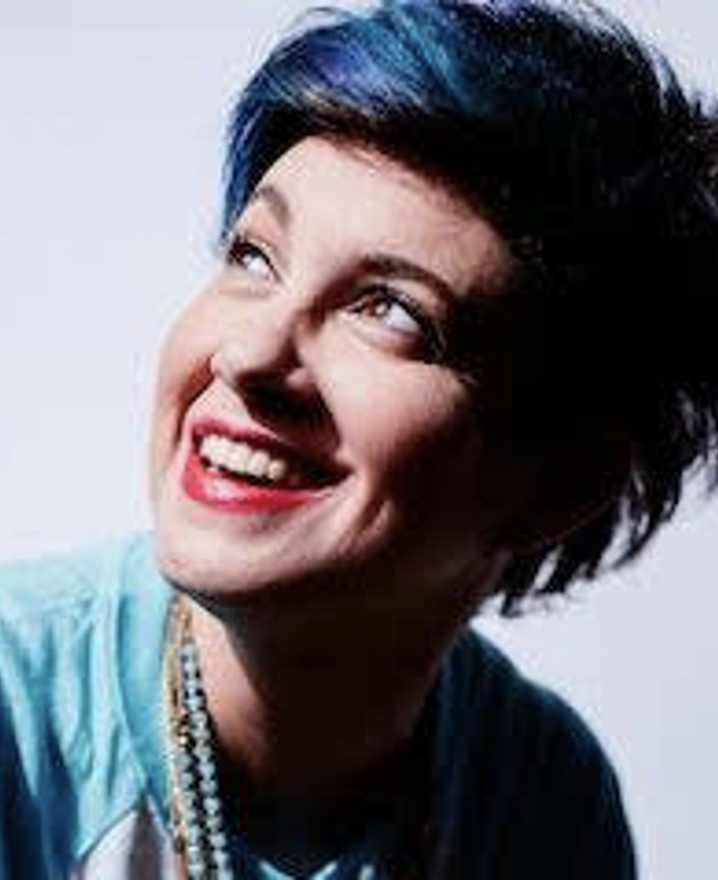 A woman with short black hair and blue highlights smiling and looking upward.