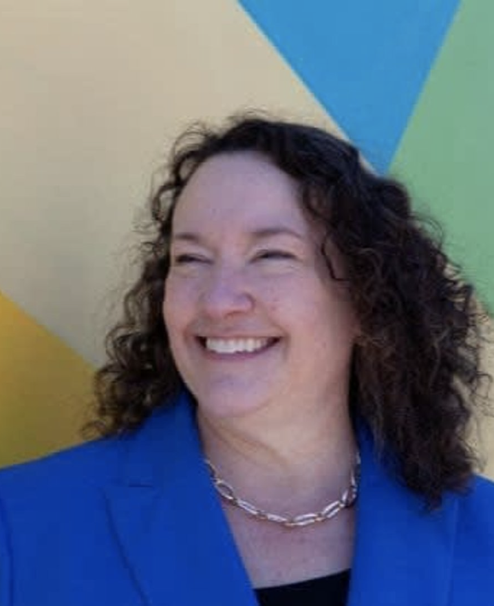 Woman with curly dark hair smiling, wearing a blue blazer and a silver chain necklace, in front of a colorful geometric background.