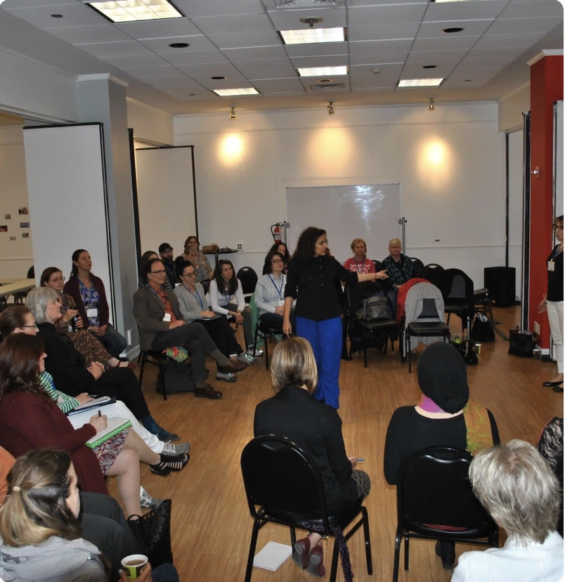 A woman standing and speaking to a group of seated women in a conference room with white walls, a whiteboard, and projection screens.