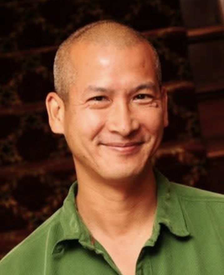 A smiling man with a shaved head wearing a green collared shirt, standing outdoors at night.