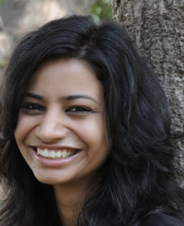 Close-up of a woman with a bright smile, dark wavy hair, standing outdoors near a tree.