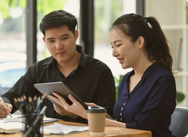 Two young adults sitting at a table, looking at a tablet together, with a coffee cup and office supplies nearby, in a bright room with large windows.