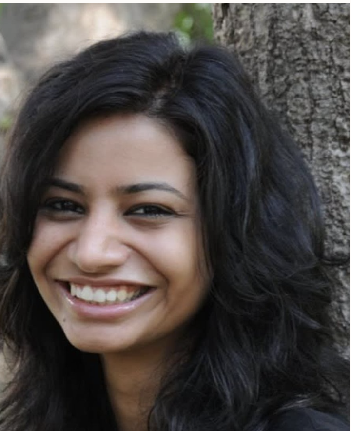 Close-up of a smiling woman with long dark hair, standing outdoors near a tree.