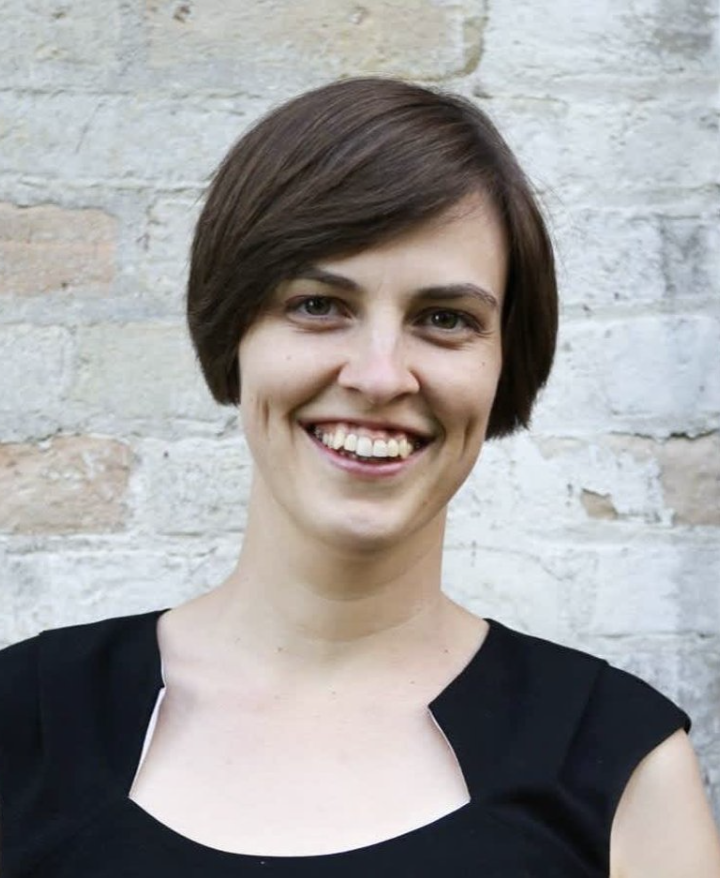 A woman with short brown hair smiling, standing against a brick wall background.