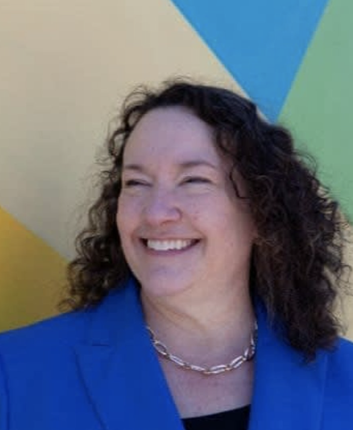 A woman with curly hair smiling, wearing a blue blazer and a silver chain, standing in front of a colorful, geometric background.