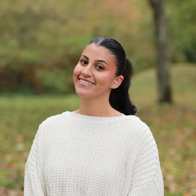 Smiling woman with dark hair tied back, wearing a white knitted sweater, outdoors in a park with green and autumn trees in the background.