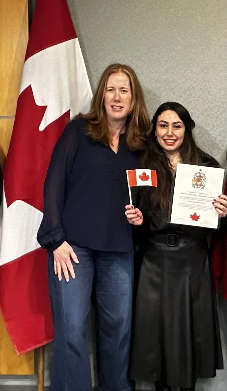 Two women standing in front of Canadian flags. One woman is holding a small Canadian flag and a certificate. They appear to be at an award or recognition ceremony.