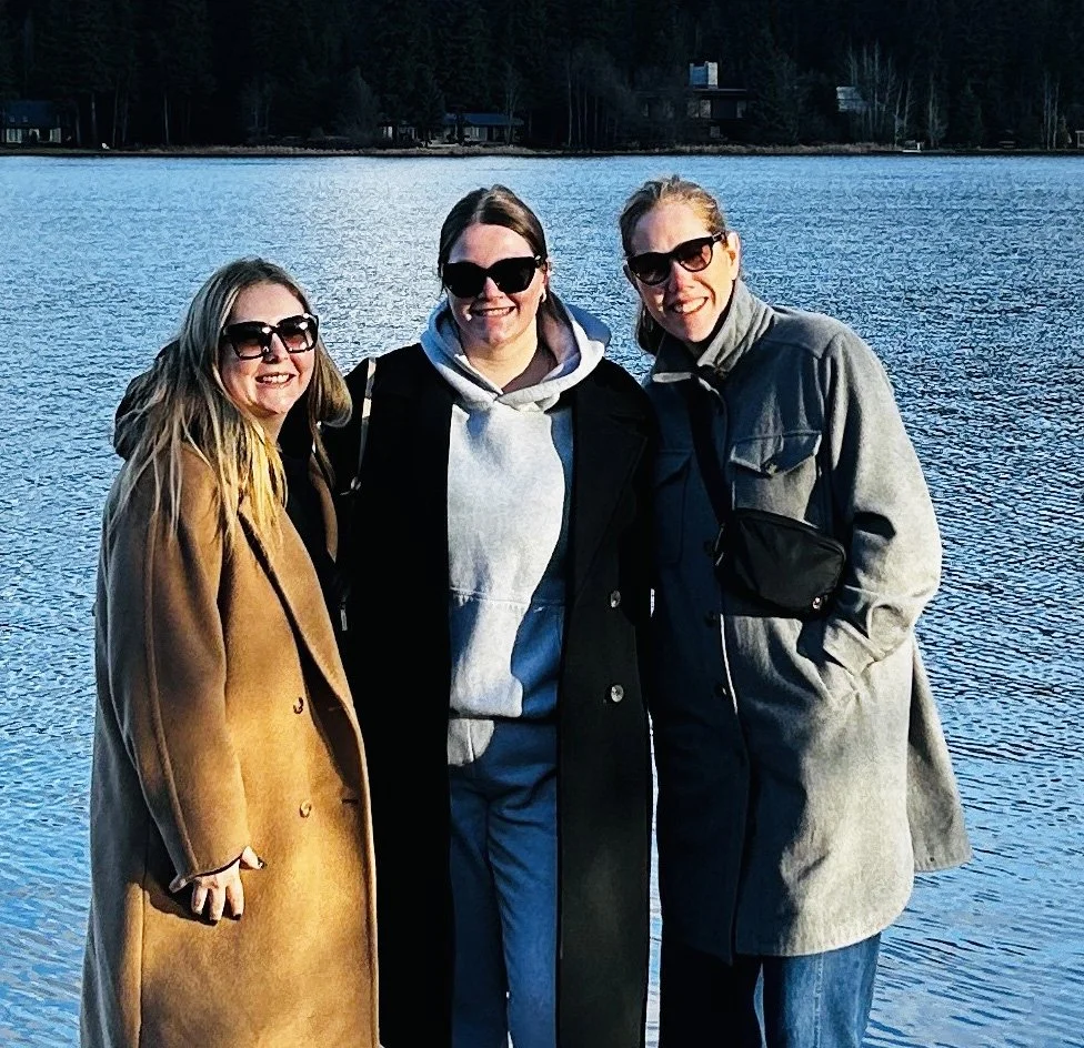 Three women in coats and sunglasses standing by a lake with trees and houses in the background.