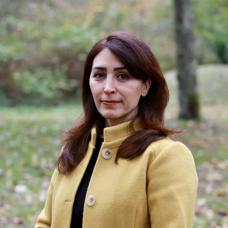 A woman with shoulder-length brown hair and dark eyebrows standing outdoors in a park with greenery and trees in the background, wearing a yellow coat with large buttons.