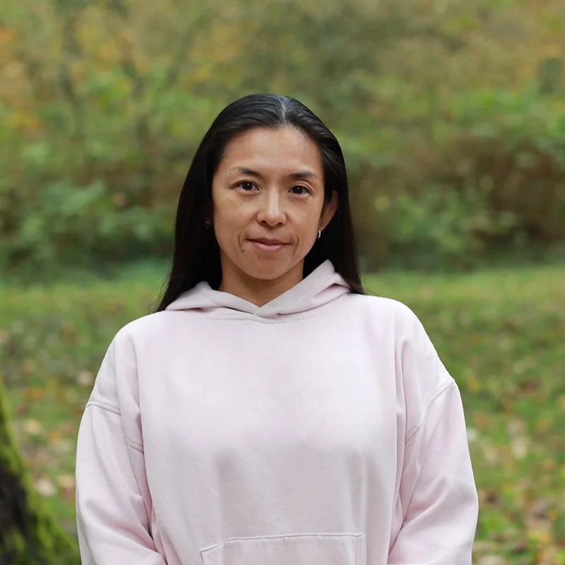 A woman with long black hair wearing a light pink hoodie standing outdoors in a forested area.