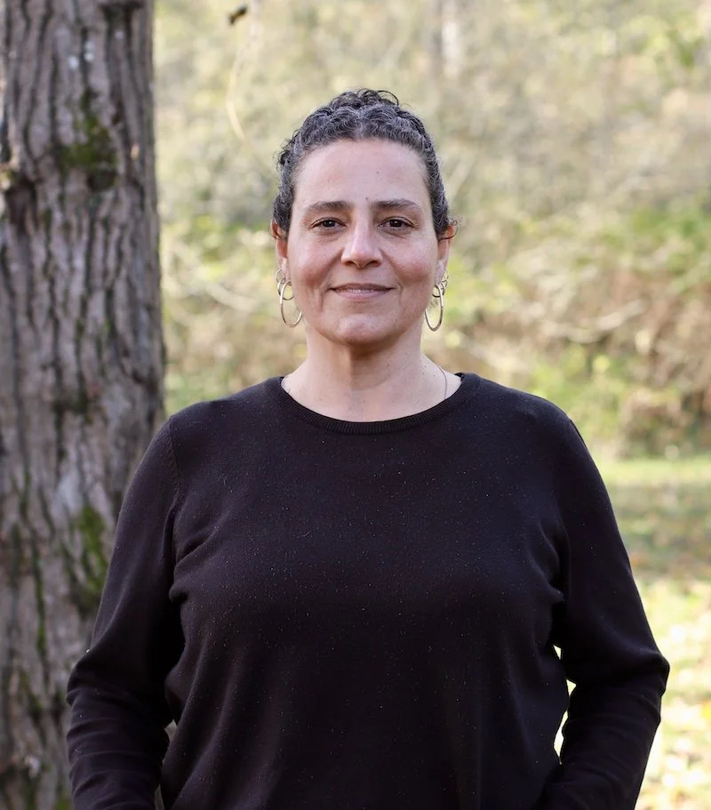 A woman with curly dark hair tied back, wearing hoops and a black sweater, is standing outdoors next to a large tree with a blurred forest background.