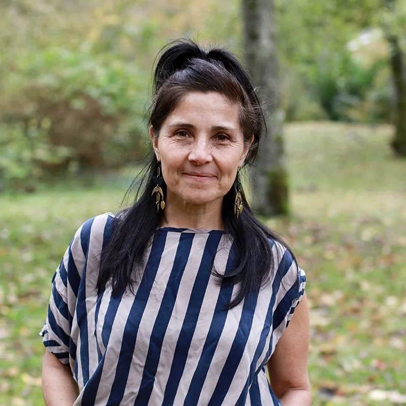 A woman with dark hair in a high ponytail, wearing a blue and beige striped top and gold earrings, standing outdoors in a wooded area with green and yellow foliage.