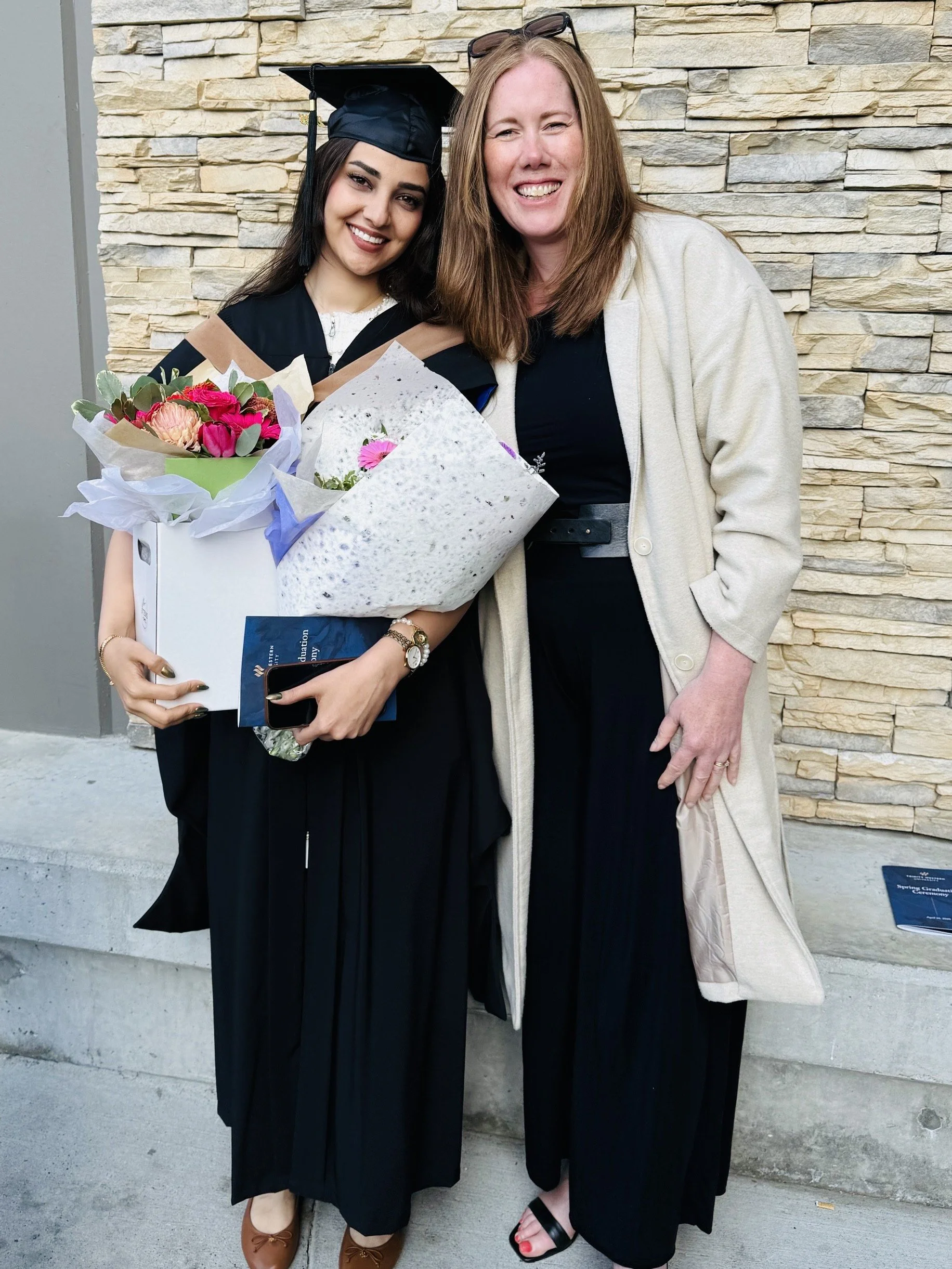 A young woman in a graduation cap and gown holding flowers and a diploma standing next to a woman in a light coat with a brick wall background.