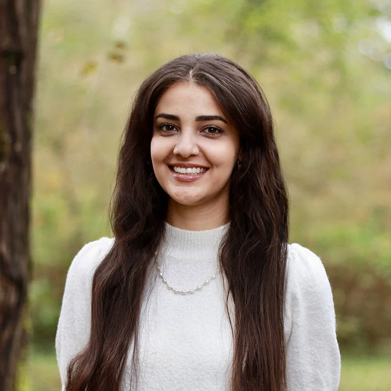A young woman with long brown hair, wearing a white sweater and necklace, smiling outdoors with green trees in the background.