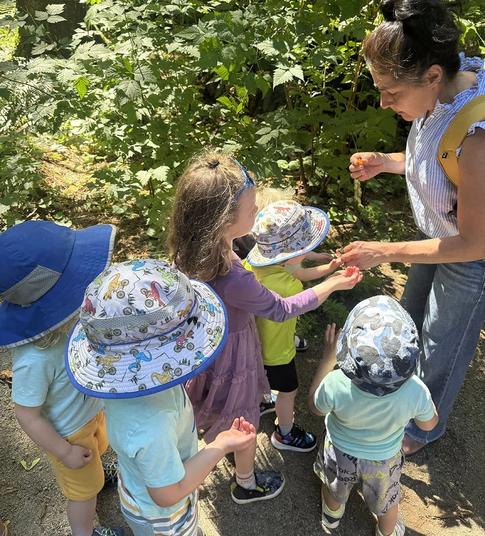 A woman and five children wearing sun hats with playful patterns are outdoors among green foliage. The woman is handing a small object to a girl in a purple dress. The children are gathered on a shaded path, engaging with the woman.