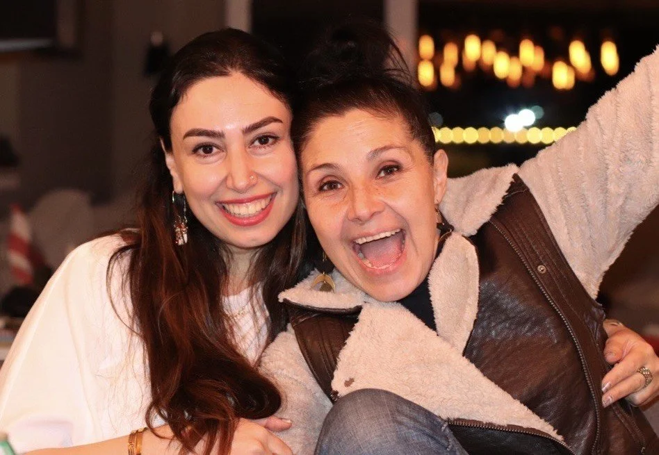 Two women smiling and taking a selfie together indoors at night, with blurred lights in the background.
