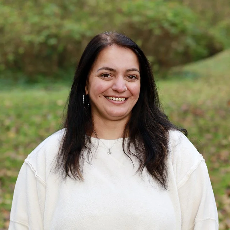 A woman with dark hair and earrings smiling outdoors in a park with green foliage.