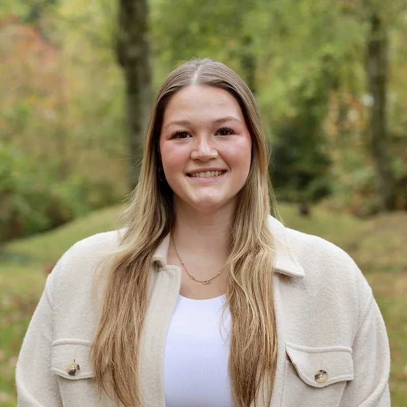 A young woman with long blonde hair smiling outdoors in a wooded area during fall, wearing a cream-colored jacket and a white top.