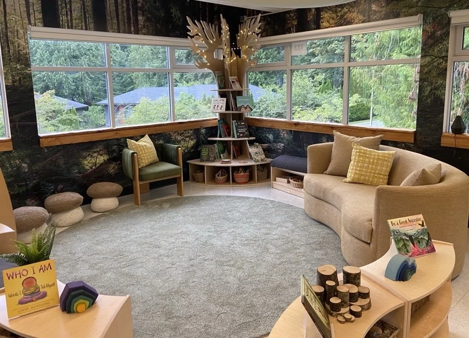 A cozy reading nook with large windows showing trees outside. There is a beige curved sofa, a green armchair, and several woven stools. In the corner, a decorative tree-shaped bookshelf with books and baskets, topped with a white sculpture of deer antlers with fairy lights. Small books and decorative items are on tables, including children's books and wooden .