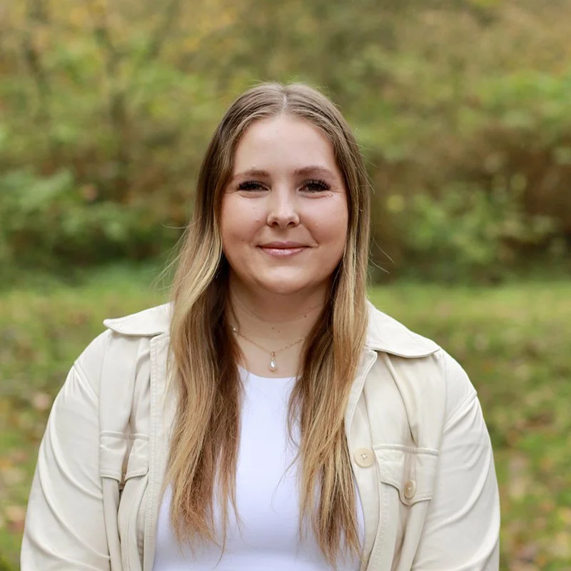 A young woman with long blonde hair, smiling, wearing a light-colored jacket and a white top, standing outdoors with autumn foliage in the background.