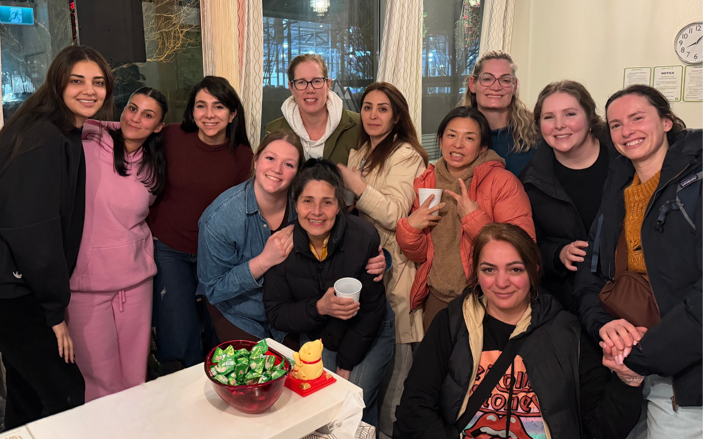 Group of women gathered together smiling indoors, with a table of snacks and drinks in front of them.