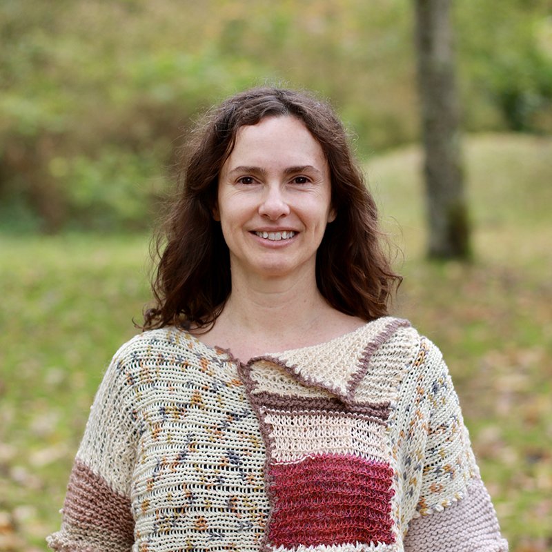 A woman with curly brown hair smiling outdoors in a park, wearing a colorful knitted sweater.