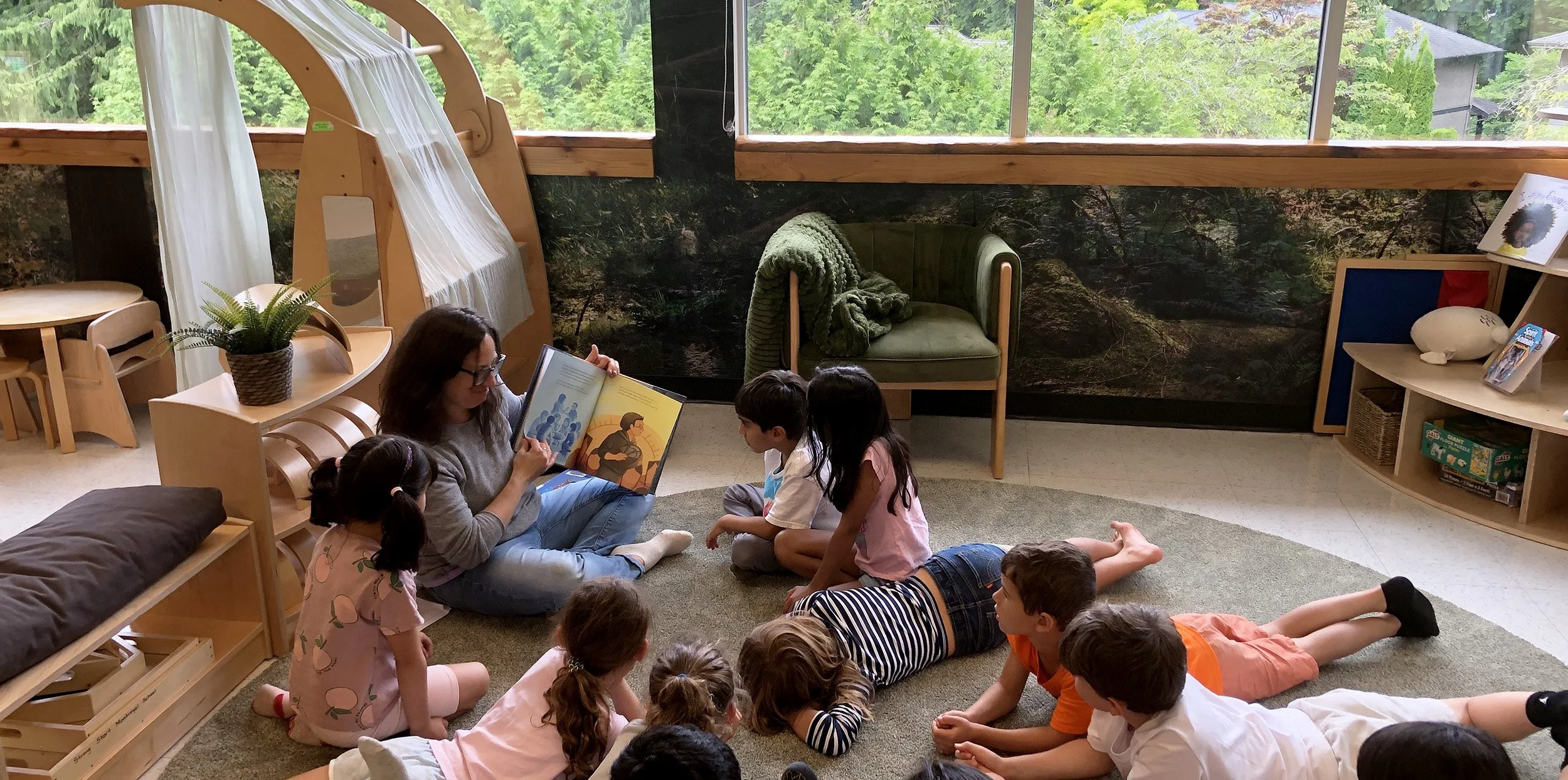 A woman reads a picture book to a group of children lying on a carpeted floor in a cozy room with large windows and a forest view.