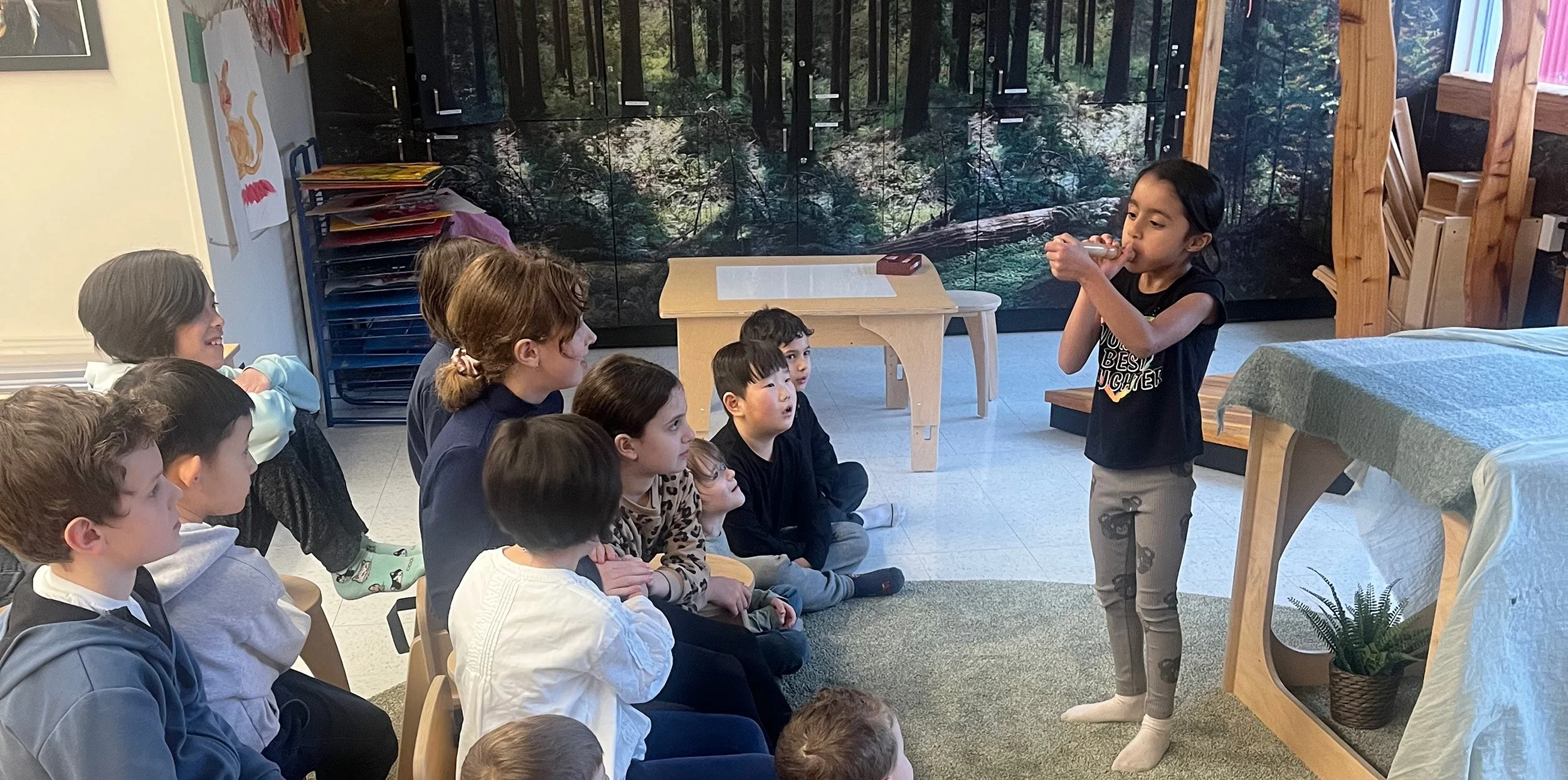 A young girl is standing and talking to a group of children who are sitting on the floor, in a classroom with a nature-themed mural in the background.